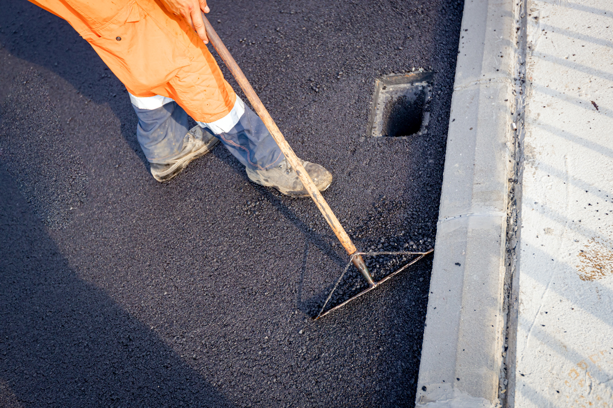 Above view on worker that uses rake to level, set up layer of fresh tarmac to right measures, flattens the hot asphalt around the quadratic rainwater hollow.